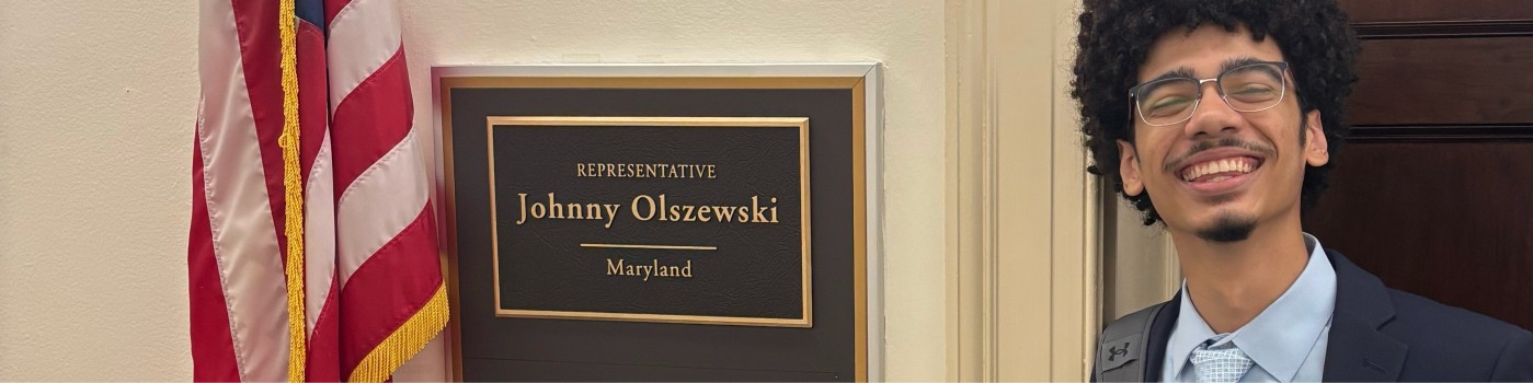 Christian Houck in a suit standing beside the brass office sign that reads 'Representative Johnny Olszewski, Maryland' on Capitol Hill, with the United States flag visible in the frame